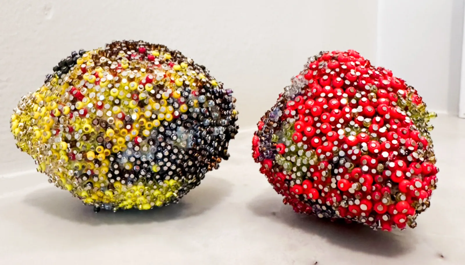 A beaded lemon and a beaded strawberry side by side on a white ledge, both encrusted edge to edge in glass seed beads and rhinestones, with dark grey and brown passages standing in for bruised skin.