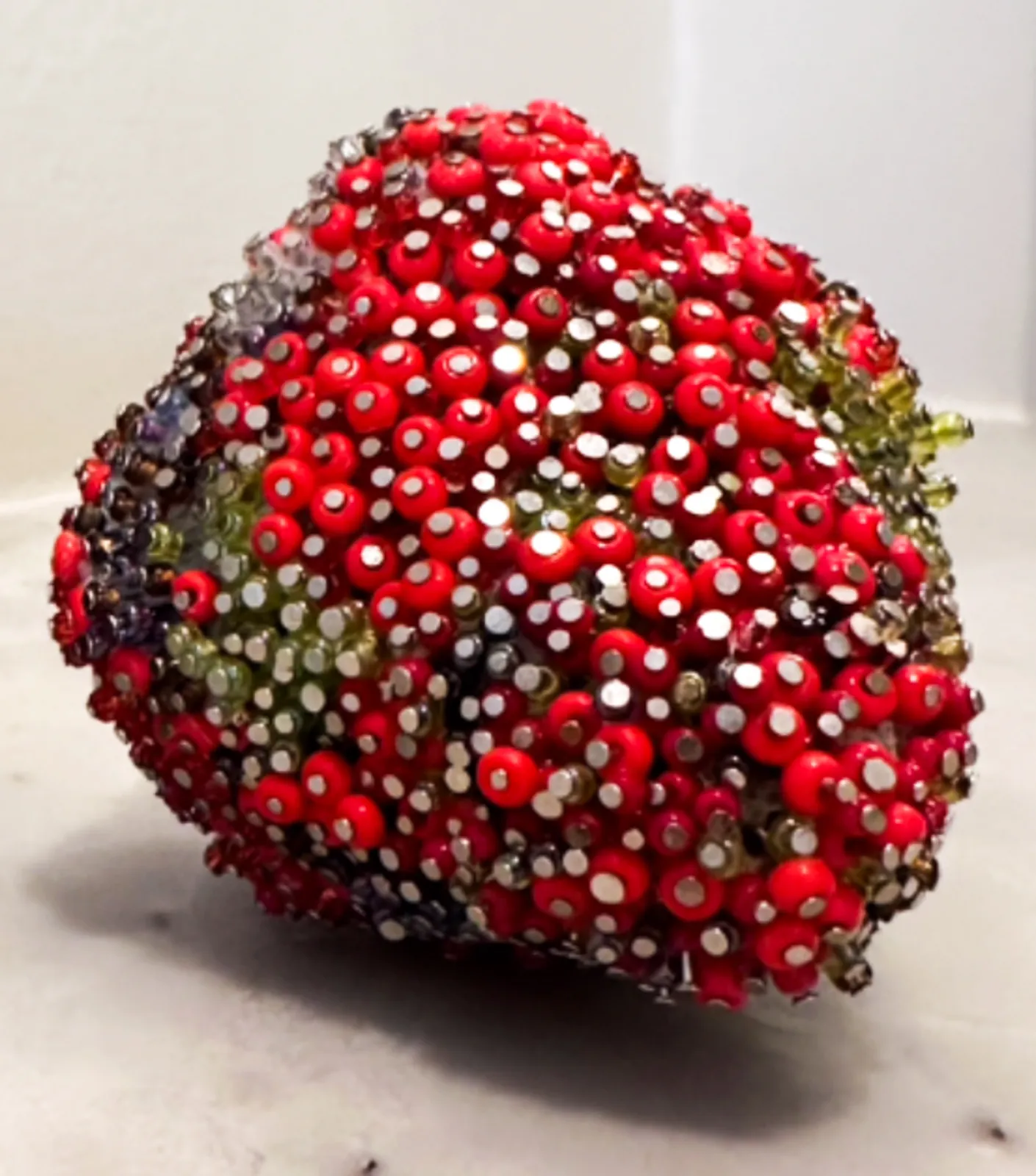 The beaded strawberry seen from above, dense red seed beads pinned with silver rhinestones and broken by patches of mold green and violet.
