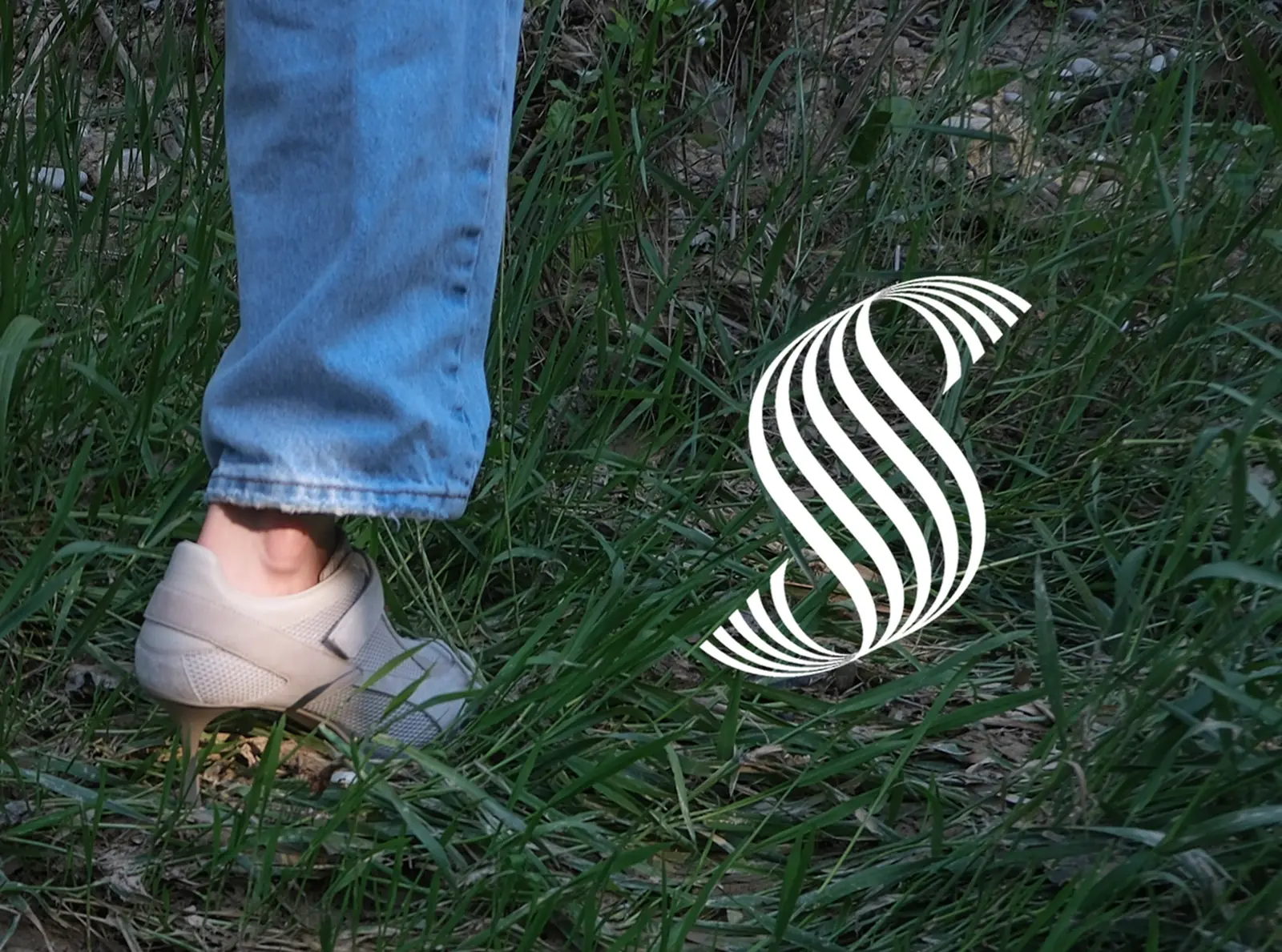 A cropped shot of one heel-soled sneaker and jeans standing in tall grass, with the Sneex spiral mark beside it.