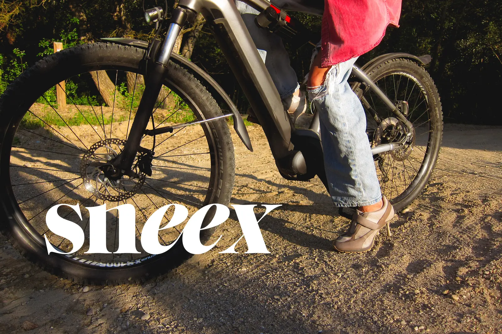 A heel-soled sneaker resting on the pedal of a mountain e-bike parked on a dirt trail, with the Sneex wordmark across the front wheel.