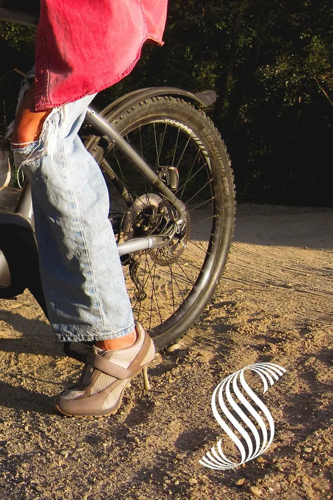 A heeled sneaker resting on the pedal of a mountain e-bike parked on a dirt trail at golden hour, with the Sneex spiral mark in the gravel behind the wheel.