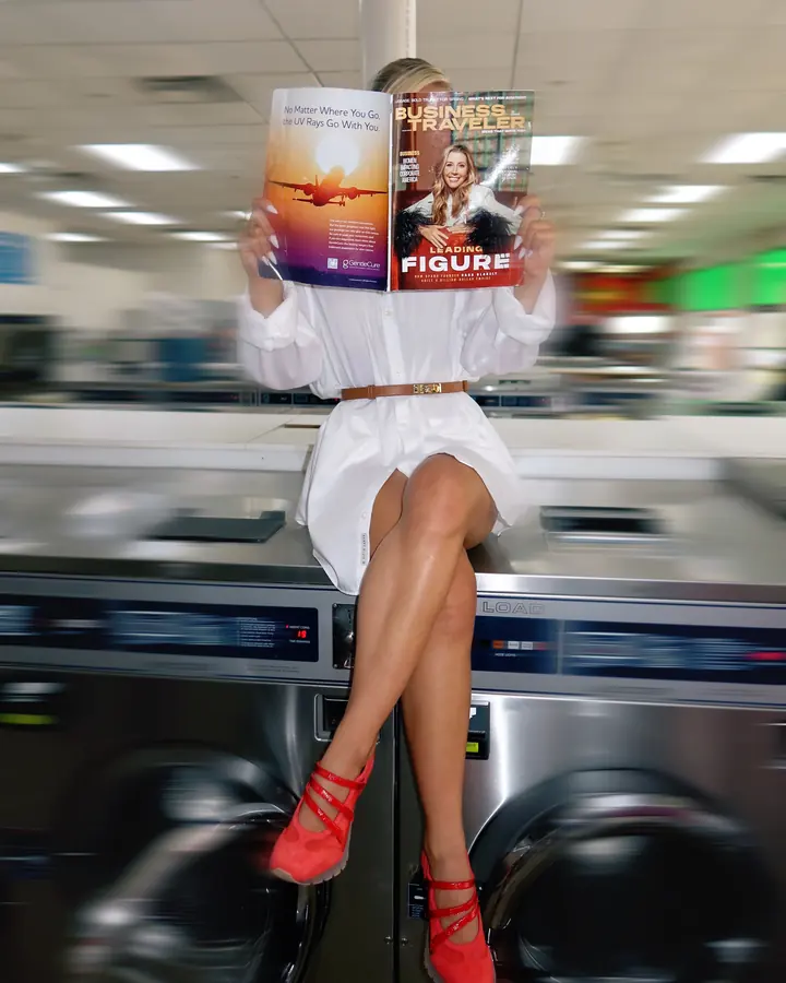 From the laundromat shoot: a model in a white shirt dress and red Sneex sits on a dryer, face hidden behind a magazine, the room blurred around her.
