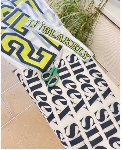 Lime BLAKELY and number transfers and rows of black Sneex wordmark transfers being cut with green-handled scissors on an ironing board.